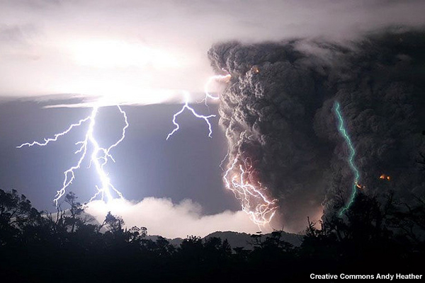 A volcano plume rises.  The plume has two lightning strikes.  A cloud a downstream produces lightning.
